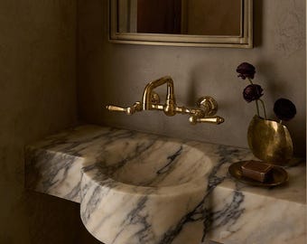 May include: A wall-mounted white marble sink with grey veining, featuring a brass faucet with two handles. Above the sink is a rectangular mirror with a curved top and two fluted white wall sconces. A small brass bowl holds a bar of soap.