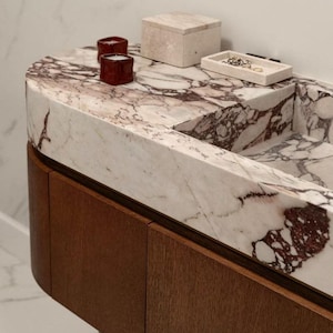 Op de afbeelding: Een moderne badkamermeubel met een marmeren aanrechtblad met bordeauxkleurige adering. Twee kleine rode kaarsen, een beige doos en een klein dienblad met sieraden staan op het aanrecht. De wastafel heeft een donkere houten kast.