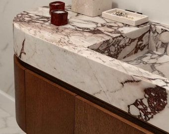Puede incluir: Un tocador de baño moderno con encimera de mármol con vetas burdeos. Dos pequeñas velas rojas, una caja beige y una pequeña bandeja con joyas se encuentran sobre la encimera. El tocador tiene un mueble de madera oscura.
