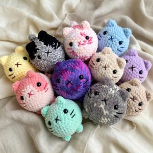 May include: A selection of crocheted cat toys in a variety of colours, such as yellow, pink, blue, and purple. Each cat has a stitched face with black eyes and a cross-stitch mouth. The toys are soft and round in shape.