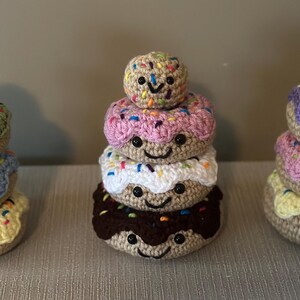 Puede incluir: Tres pilas de donas de crochet con caras sonrientes. Cada pila tiene cuatro donas, con la dona superior siendo una galleta más pequeña. Las donas están decoradas con chispas de colores y glaseado en diferentes colores, incluyendo rosa, morado, verde, azul, blanco y marrón.