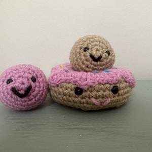 May include: Three crocheted stuffed toys with smiling faces. The largest toy is a donut with pink frosting and sprinkles. A smaller brown ball sits on top of the donut. A smaller pink ball sits to the left of the donut.