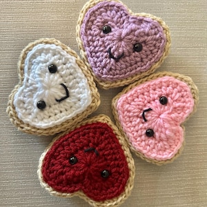 Può includere: Quattro panini a forma di cuore all'uncinetto con ripieni di diversi colori. I panini sono bianchi, rosa, viola e rossi. Ogni panino ha un viso sorridente con occhi neri.