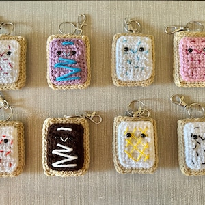 May include: Eight crocheted keychains shaped like pop-tart pastries with different colored fillings and faces. The keychains are on a light brown surface.