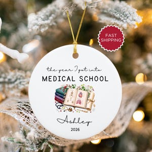 Op de afbeelding: Witte keramische kerstversiering met een gouden touwtje. De versiering leest "the year I got into MEDICAL SCHOOL" met een medische illustratie en de naam "Ashley" en het jaar "2026".