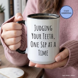 May include: A white ceramic mug with a black handle and rim, filled with coffee. The mug has the text "JUDGING YOUR TEETH, ONE SIP AT A TIME" in black. The mug is held by a person wearing a pink sweater. The mug is 425ml.