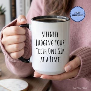 May include: A white ceramic mug with a black rim and handle, filled with coffee. The mug features the text "SILENTLY JUDGING YOUR TEETH ONE SIP AT A TIME" in black. A blue "FAST SHIPPING" sticker is visible.
