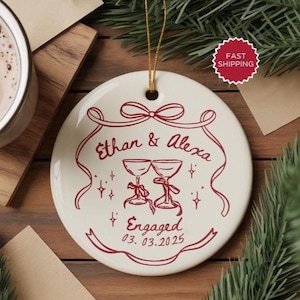May include: A round, off-white ceramic ornament with a red design. The ornament features the names "Ethan & Alexa", two champagne glasses, the word "Engaged", and the date 03.03.2025. A gold string is attached for hanging.