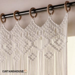 以下が含まれることがあります： 幾何学模様の白いマクラメカーテン。木製のリングが付いた濃い色の金属棒から吊り下げられています。カーテンはスカラップのトップエッジと長いフリンジが特徴です。「CURTAINSHOUSE」の文字が左下に表示されています。