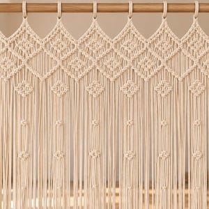 May include: A cream-coloured macrame curtain hanging from a wooden rod with circular rings. The top section features a repeating diamond pattern created with intricate knots, while the lower section consists of long, vertical hanging cords.