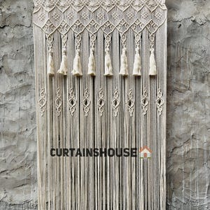 Puede incluir: Una cortina de macram&eacute; color hueso colgada de una barra de metal, con intrincados patrones geom&eacute;tricos anudados en la parte superior, una fila de borlas colgantes y una serie de peque&ntilde;os nudos en forma de diamante. El texto CURTAINHOUSE es visible.