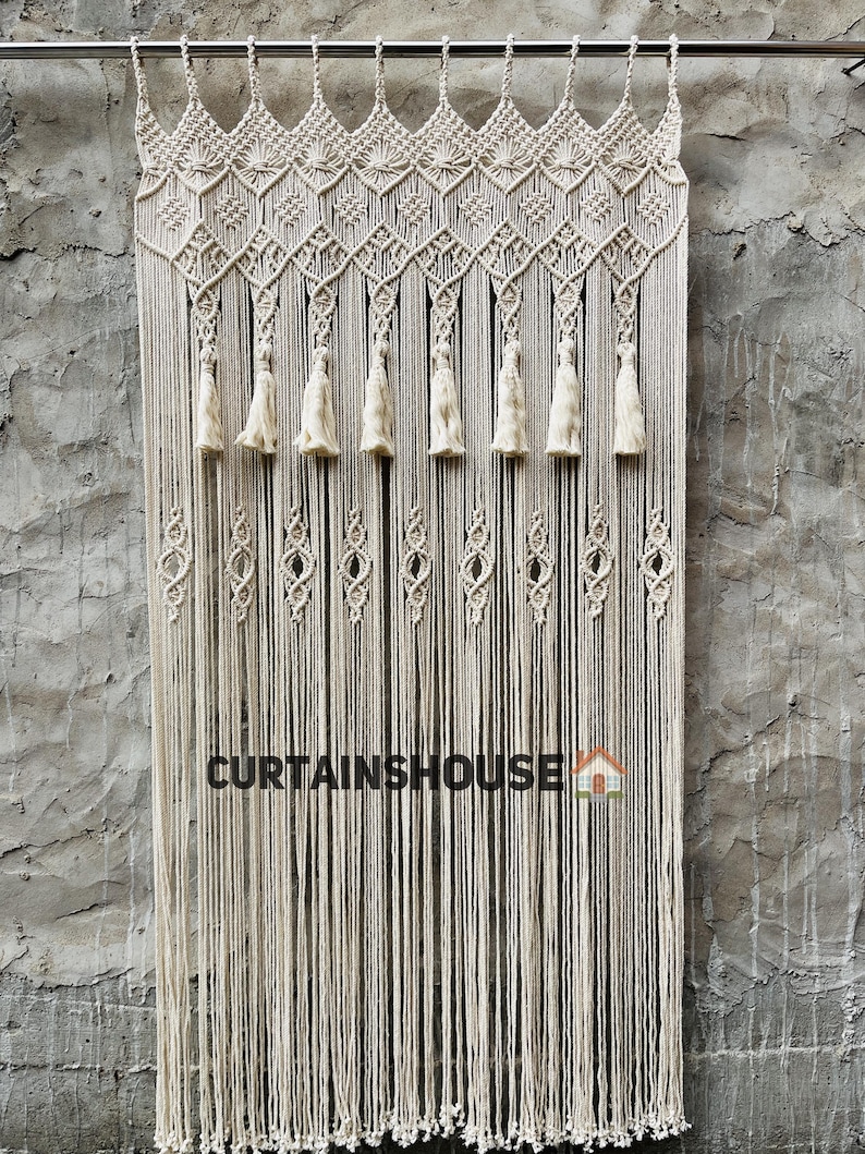 Puede incluir: Una cortina de macram&eacute; color hueso colgada de una barra de metal, con intrincados patrones geom&eacute;tricos anudados en la parte superior, una fila de borlas colgantes y una serie de peque&ntilde;os nudos en forma de diamante. El texto CURTAINHOUSE es visible.