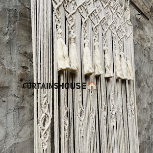 Puede incluir: Un tapiz de macram&eacute; de color crema con intrincados patrones de nudos, dise&ntilde;os en forma de diamante y borlas colgantes suspendidas de una barra de metal. El texto CURTAINSHOUSE con un peque&ntilde;o icono de casa est&aacute; impreso en la parte frontal.
