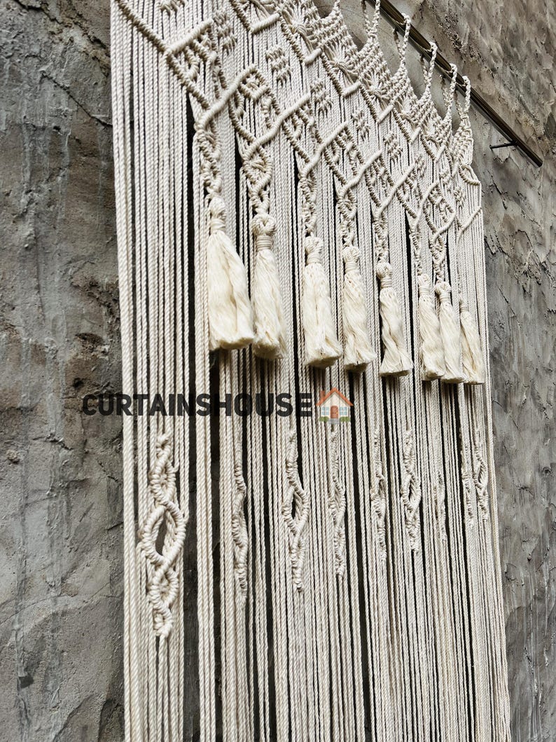 Puede incluir: Un tapiz de macram&eacute; de color crema con intrincados patrones de nudos, dise&ntilde;os en forma de diamante y borlas colgantes suspendidas de una barra de metal. El texto CURTAINSHOUSE con un peque&ntilde;o icono de casa est&aacute; impreso en la parte frontal.