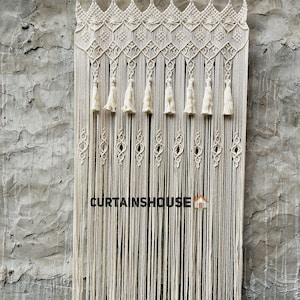 Puede incluir: Una cortina de macram&eacute; color hueso colgada de una barra de metal, con patrones de nudos complejos en la parte superior, una fila de borlas decorativas y largos flecos verticales. El texto CURTAINSHOUSE est&aacute; impreso en negro con un icono de casa.