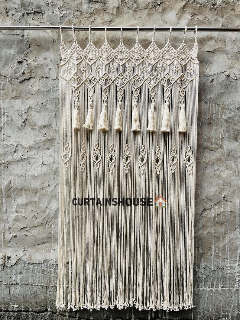 Puede incluir: Una cortina de macram&eacute; color hueso colgada de una barra de metal, con patrones de nudos complejos en la parte superior, una fila de borlas decorativas y largos flecos verticales. El texto CURTAINSHOUSE est&aacute; impreso en negro con un icono de casa.