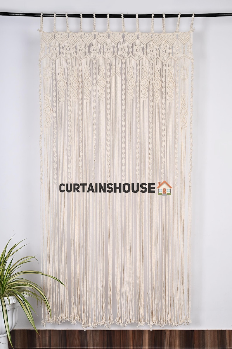 K&ouml;nnte beinhalten: Ein wei&szlig;er Makramee-Vorhang mit geometrischem Muster. Der Vorhang h&auml;ngt an einer schwarzen Stange und ist aus Baumwollschnur gefertigt. Der Text "CURTAINS HOUSE" ist unten in der Mitte des Bildes sichtbar.
