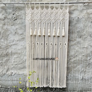 Puede incluir: Una cortina de macram&eacute; color hueso colgada de una barra met&aacute;lica, con un intrincado patr&oacute;n de rombos anudados en la parte superior, una fila de ocho borlas y largos flecos verticales. El texto CURTAINHOUSE est&aacute; impreso en negro en la parte inferior.