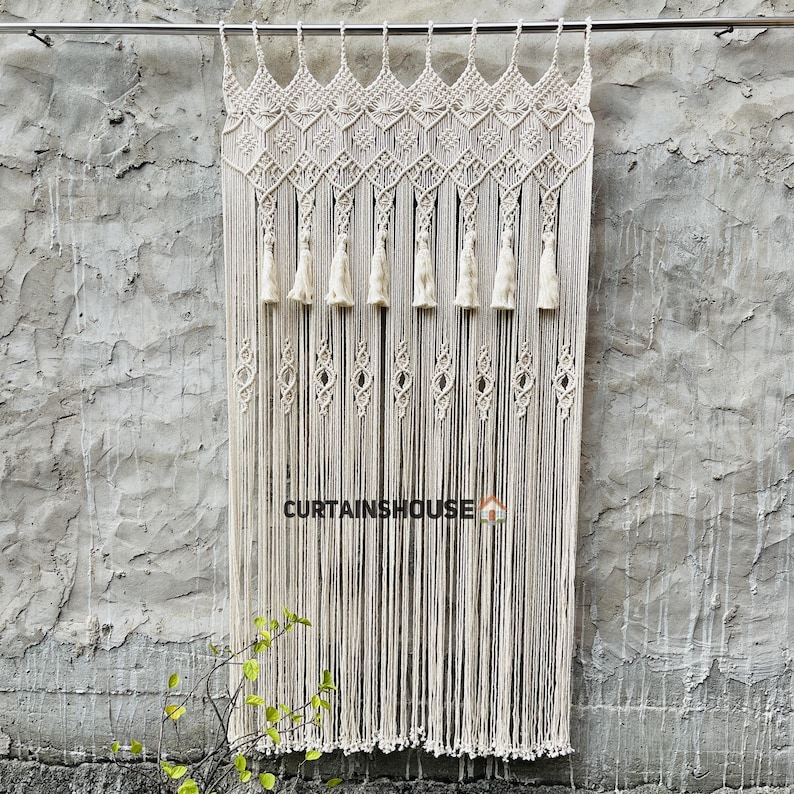 Puede incluir: Una cortina de macram&eacute; color hueso colgada de una barra met&aacute;lica, con un intrincado patr&oacute;n de rombos anudados en la parte superior, una fila de ocho borlas y largos flecos verticales. El texto CURTAINHOUSE est&aacute; impreso en negro en la parte inferior.