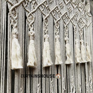 Puede incluir: Un tapiz de macram&eacute; color crema con intrincados patrones de nudos, dise&ntilde;os en forma de diamante y una serie de borlas colgantes en la parte inferior. El texto CURTAINSHOUSE est&aacute; impreso en negro cerca del centro inferior de la pieza de fibra.