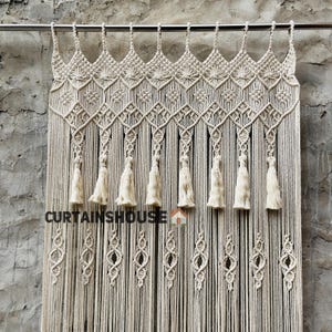 Puede incluir: Una cortina de macram&eacute; de color crema colgada de una barra de metal, con intrincados patrones de nudos, una serie de borlas colgantes y un dise&ntilde;o de rombos repetido. El texto CURTAINHOUSE est&aacute; impreso en negro en el centro de la cortina.