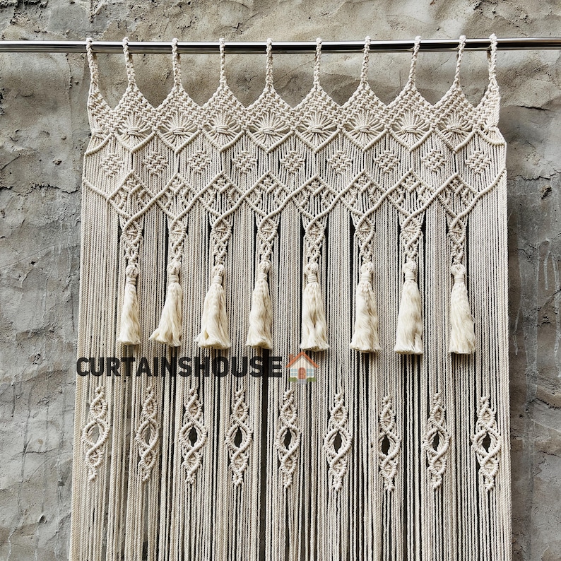 Puede incluir: Una cortina de macram&eacute; de color crema colgada de una barra de metal, con intrincados patrones de nudos, una serie de borlas colgantes y un dise&ntilde;o de rombos repetido. El texto CURTAINHOUSE est&aacute; impreso en negro en el centro de la cortina.