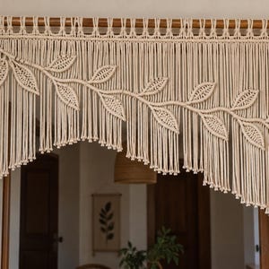 May include: An off-white macrame curtain hanging from a wooden rod, featuring a central diagonal branch design with multiple woven leaf-shaped elements and long, vertical fringe strands that create a layered, asymmetrical bottom edge.