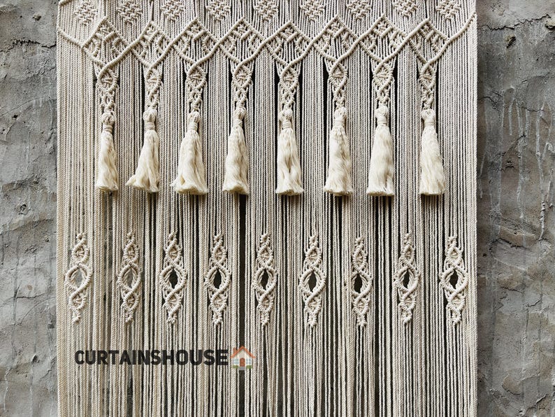 Puede incluir: Un tapiz de macram&eacute; color crema con intrincados patrones de nudos, dise&ntilde;os geom&eacute;tricos en forma de diamante y una fila de borlas colgantes. La secci&oacute;n inferior muestra hilos verticales repetidos y el texto CURTAINHOUSE est&aacute; impreso en negro en la parte inferior izquierda.