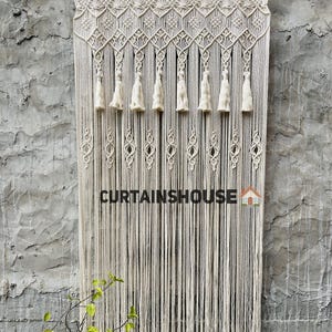 Puede incluir: Una cortina de macram&eacute; de color crema que presenta una secci&oacute;n superior anudada intrincada con patrones geom&eacute;tricos, borlas colgantes y largos hilos de flecos verticales. El texto CURTAINSHOUSE est&aacute; impreso en negro con un peque&ntilde;o icono de casa.