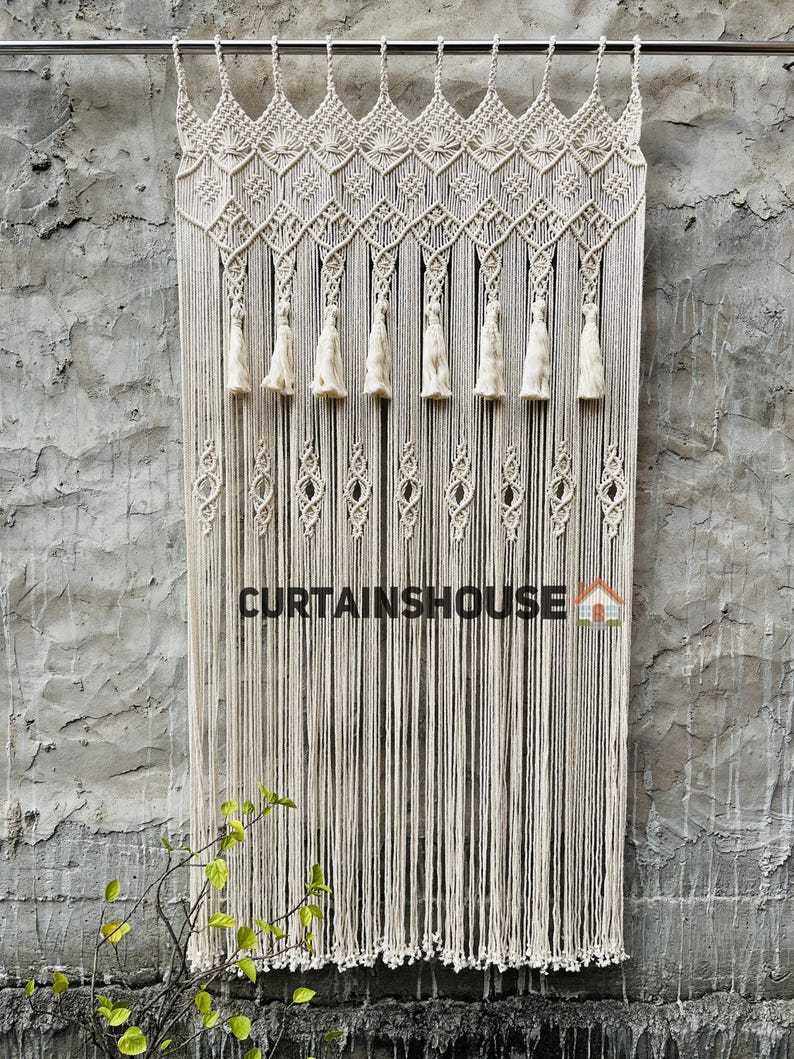 Puede incluir: Una cortina de macram&eacute; de color crema que presenta una secci&oacute;n superior anudada intrincada con patrones geom&eacute;tricos, borlas colgantes y largos hilos de flecos verticales. El texto CURTAINSHOUSE est&aacute; impreso en negro con un peque&ntilde;o icono de casa.