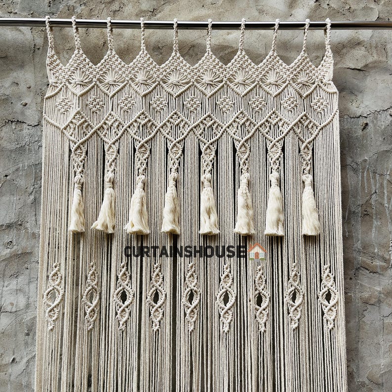 Puede incluir: Una cortina de macram&eacute; color hueso colgada de una barra de metal, con patrones de nudos intrincados, un borde superior en forma de diamante y ocho borlas colgantes. El texto CURTAINSHOUSE con un peque&ntilde;o icono de casa est&aacute; impreso en la cortina.