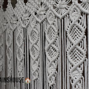 May include: White macrame curtain with intricate woven patterns. The design features diamond and floral motifs, creating a textured, bohemian aesthetic. The curtain is made of thick, woven cotton rope, and is suitable for a window or doorway.