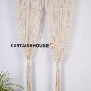 K&ouml;nnte beinhalten: Zwei cremefarbene Makramee-Vorh&auml;nge mit geometrischem Muster. Die Vorh&auml;nge h&auml;ngen an einer schwarzen Stange und haben lange Quasten am unteren Ende. Der Text "CURTAINS HOUSE" ist im Bild sichtbar.