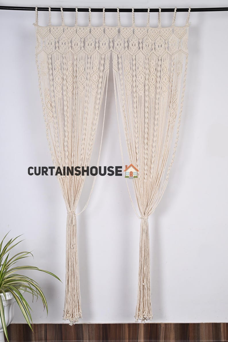 K&ouml;nnte beinhalten: Zwei cremefarbene Makramee-Vorh&auml;nge mit geometrischem Muster. Die Vorh&auml;nge h&auml;ngen an einer schwarzen Stange und haben lange Quasten am unteren Ende. Der Text "CURTAINS HOUSE" ist im Bild sichtbar.