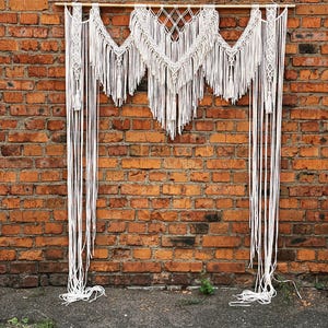May include: A white macrame wall hanging with a geometric design and long fringe. The macrame is suspended from a wooden dowel and hangs against a red brick wall. The piece has a bohemian aesthetic.