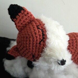 May include: A close-up of a handmade fox head. The fox has a crocheted orange head with black accents and a white fluffy chest. The fox's ear is orange with a black tip.