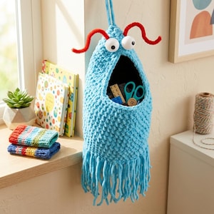May include: A whimsical, light blue, crocheted storage basket shaped like a monster with googly eyes and red pipe cleaner antennae. The basket has a fringed bottom and is hanging. Inside are scissors and other small items. A stack of colorful crocheted coasters sits on a windowsill.