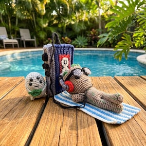 May include: A collection of handmade crochet items, including a gray creature with a green scarf, a blue and gray cassette tape-shaped bag, and a brown creature wearing sunglasses and headphones, relaxing on a blue and white striped towel.