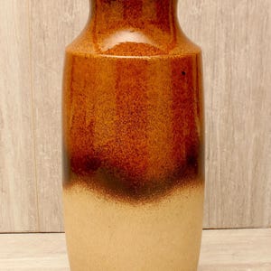 Könnte beinhalten: Eine hohe Keramikvase mit zylindrischem Körper und ausgestelltem Rand. Der obere Teil der Vase ist mit einer glänzenden, tief rotbraunen Glasur versehen, die in einen matten, hellbeigen Farbton im unteren Bereich des Gefäßes übergeht.