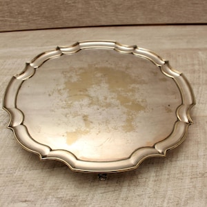 Op de afbeelding: Een rond zilverkleurig dienblad met een geschulpte rand en een glad, reflecterend oppervlak. De metalen afwerking vertoont tekenen van slijtage en aanslag, vooral in het midden, met een gepolijste rand die het licht opvangt.