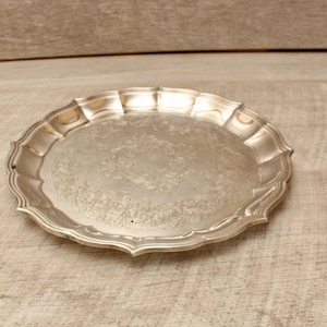 Puede incluir: Una bandeja redonda de metal plateado con un borde festoneado y un patrón floral en relieve decorativo centrado en la superficie plana. La bandeja tiene un acabado pulido y reflectante, con un borde elevado que sigue el contorno curvo.