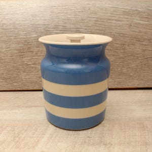 May include: A ceramic canister featuring a glossy finish with alternating horizontal stripes in blue and off-white. The container has a wide, flared rim and a matching lid with a small, centred knob handle on top for easy removal and secure closure.