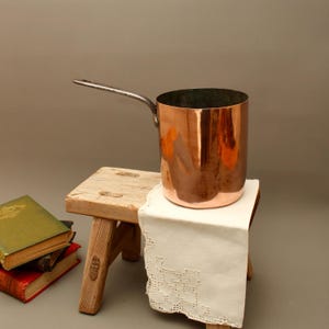 Pode incluir: Uma panela de cobre polido com um cabo longo de metal escuro. A panela apresenta um corpo cilíndrico com uma superfície reflexiva e um interior escuro. Está posicionada sobre um pequeno banco de madeira coberto com um pano branco com renda.
