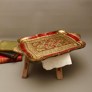 Puede incluir: Una bandeja de servicio rectangular con acabado en oro y rojo, que presenta un patrón en relieve ornamentado con pequeños detalles en azul y rojo. La bandeja se apoya sobre un pequeño taburete de madera con un tapete de encaje.