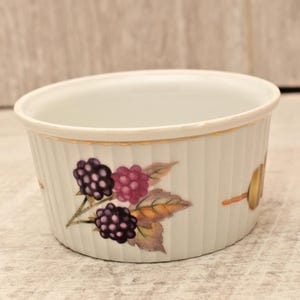May include: A white porcelain ramekin featuring a fluted exterior design with a gold-trimmed rim. The side of the dish is decorated with a botanical illustration of dark purple and pink blackberries accompanied by green and brown leaves.