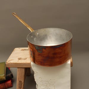 May include: A round copper saucepan with a metallic interior and a long, angled brass handle. The exterior of the pan displays a warm, polished copper finish with visible areas of natural oxidation and dark patina, showing a cylindrical shape.