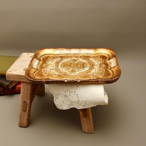 May include: A rectangular gold-toned tray with an ornate embossed pattern rests on a small, light-coloured wooden stool. A white lace doily with a delicate floral cutout design hangs over the front edge of the stool, partially covering the legs.