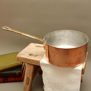 May include: A round copper saucepan with a long, polished brass handle. The exterior of the pan features a warm, metallic copper finish with subtle surface variations, while the interior is lined with a smooth, silver-toned metallic material.