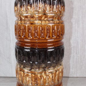 Può includere: Un vaso cilindrico in ceramica con finitura lucida e bande alternate di smalto marrone scuro e color miele. La superficie è caratterizzata da una texture con un motivo rettangolare verticale ripetuto che avvolge il corpo del contenitore.