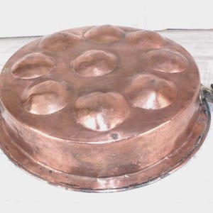 May include: A round copper pan featuring a series of nine circular indentations on its top surface. The pan has a flat rim and two dark metal handles attached to opposite sides. The copper surface shows a warm, metallic, reddish-brown finish.